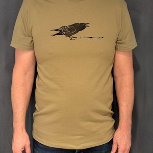 Peut inclure: T-shirt vert olive avec un dessin noir d'un oiseau au bec ouvert. L'oiseau est sur une ligne. Le t-shirt a des manches courtes et un col rond.