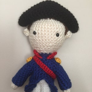 May include: A crocheted doll wearing a blue and red uniform with a black hat. The doll has white skin, black eyes, and a red mouth.