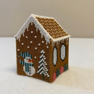 May include: A small gingerbread house cross-stitch featuring a snowman, a tree, and candy canes. The house is brown with white icing and gold glitter on the roof. The snowman is wearing a blue and green hat and scarf.