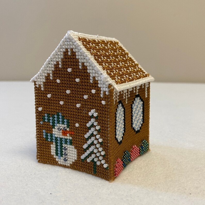 May include: A small gingerbread house cross-stitch featuring a snowman, a tree, and candy canes. The house is brown with white icing and gold glitter on the roof. The snowman is wearing a blue and green hat and scarf.