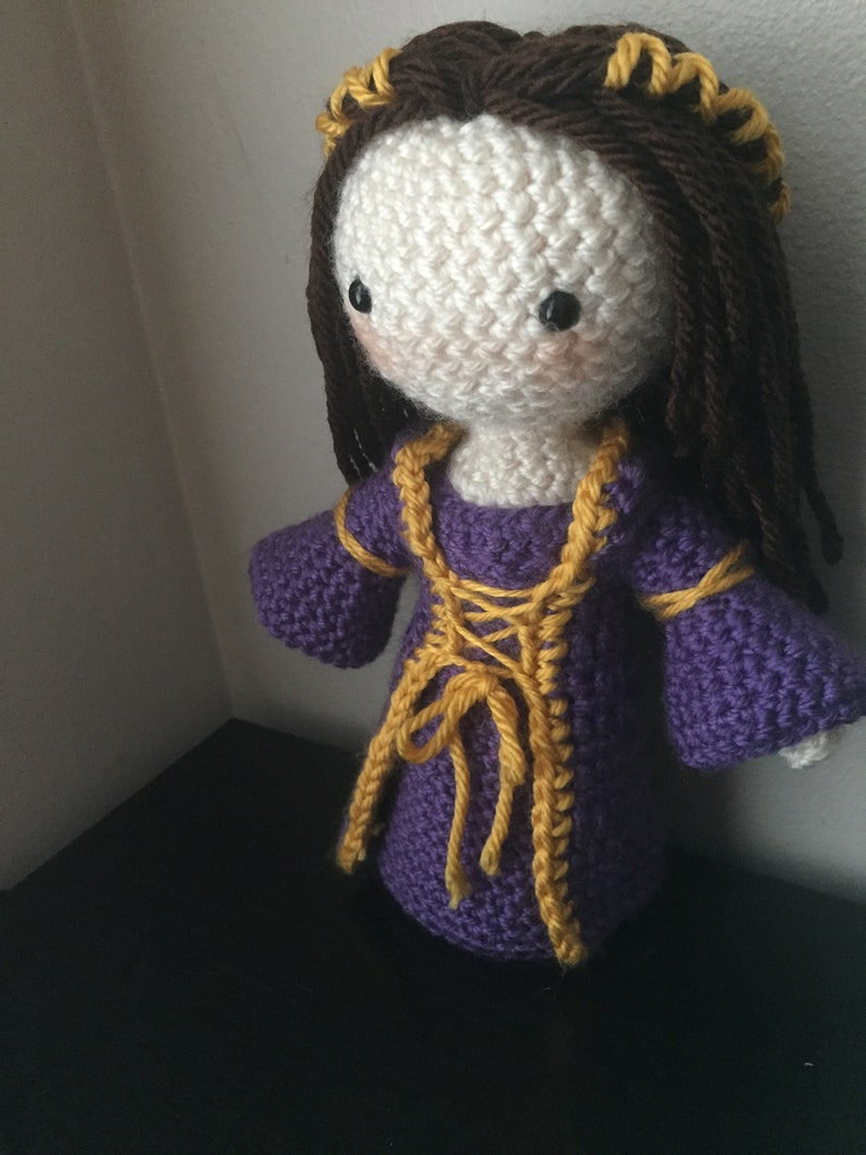 May include: Crocheted doll with brown hair, a purple dress with yellow trim, and a yellow crown.