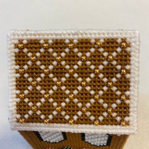 May include: A small gingerbread house with a white roof decorated with gold beads. The house has white windows and a brown base with colourful beads.