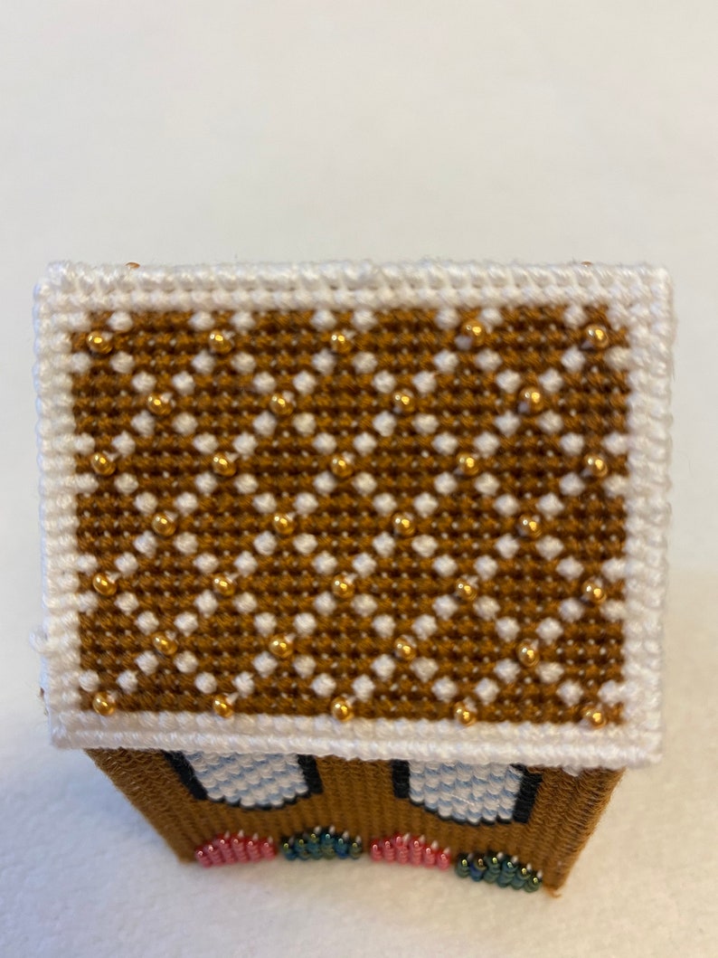 May include: A small gingerbread house with a white roof decorated with gold beads. The house has white windows and a brown base with colourful beads.