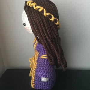 May include: Crocheted doll with brown hair and a yellow crown, wearing a purple and yellow dress.