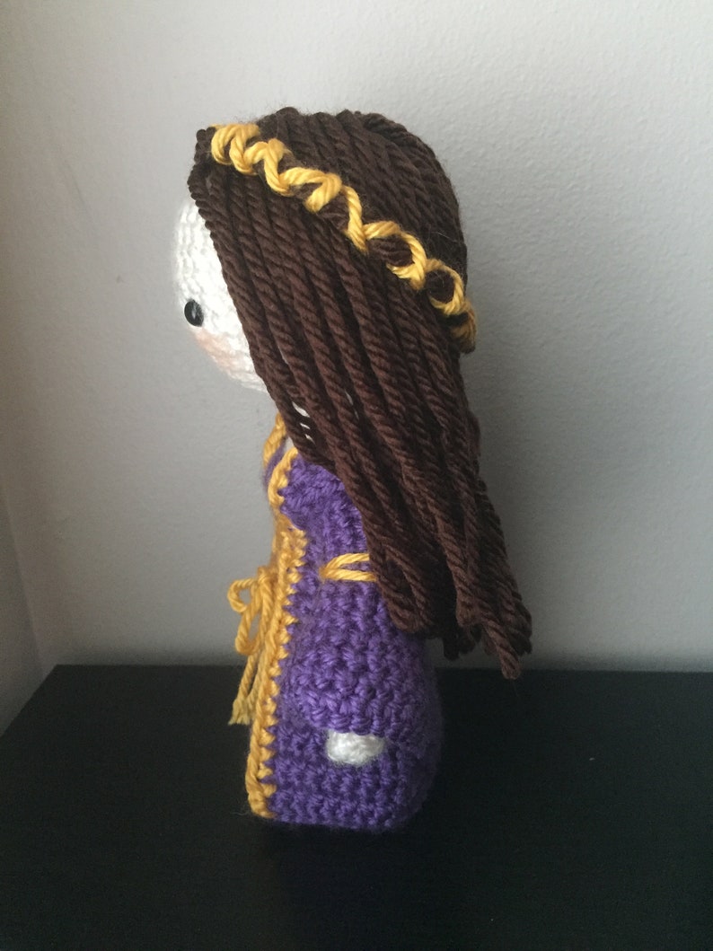 May include: Crocheted doll with brown hair and a yellow crown, wearing a purple and yellow dress.