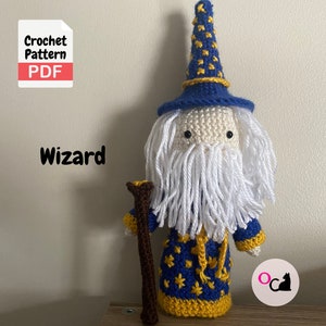 Puede incluir: Patrón de crochet PDF para un amigurumi de mago. La muñeca lleva una túnica azul y amarilla con estrellas, una barba blanca y un sombrero puntiagudo azul con estrellas amarillas. La muñeca sostiene un palo marrón.