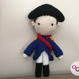 May include: A crocheted doll wearing a blue jacket, white pants, a red sash, and a black hat. The doll has a white face with black eyes and a yellow button nose.