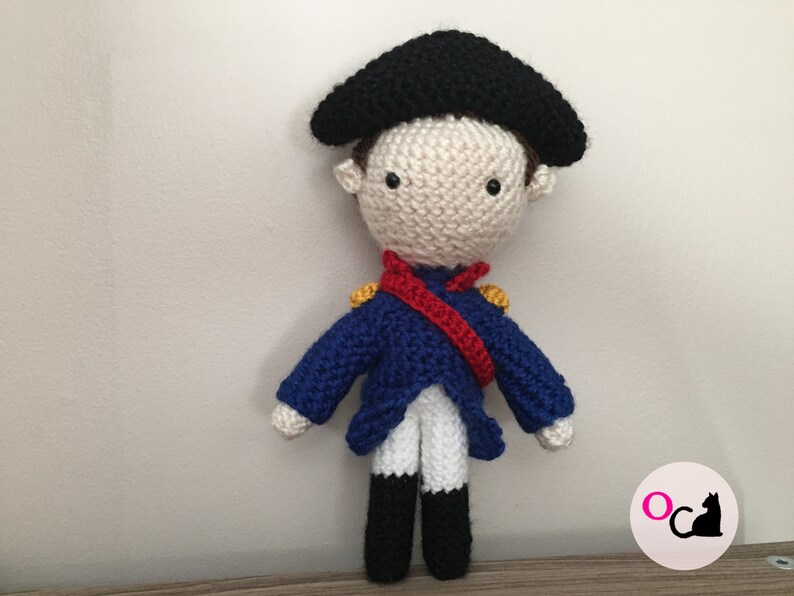 May include: A crocheted doll wearing a blue jacket, white pants, a red sash, and a black hat. The doll has a white face with black eyes and a yellow button nose.