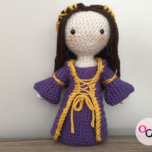 May include: A crocheted doll wearing a purple dress with yellow trim and a brown wig. The doll has a white face with black eyes and pink cheeks.