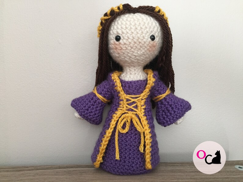 May include: A crocheted doll wearing a purple dress with yellow trim and a brown wig. The doll has a white face with black eyes and pink cheeks.
