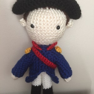May include: A crocheted doll wearing a blue jacket, white pants, black shoes, and a black hat. The doll has a red sash and gold accents on the jacket.