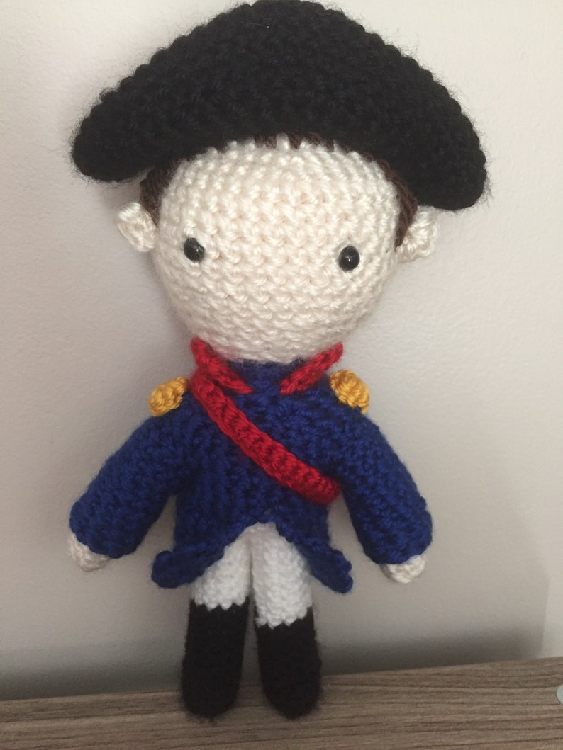 May include: A crocheted doll wearing a blue jacket, white pants, black shoes, and a black hat. The doll has a red sash and gold accents on the jacket.