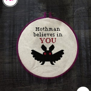Puede incluir: Un aro de bordado blanco con un borde morado, que presenta un murciélago negro con ojos rojos y el texto "Mothman believes in YOU".