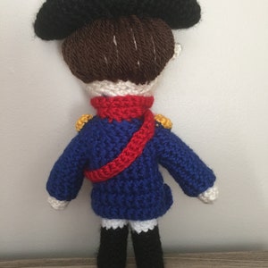 May include: A crocheted doll wearing a blue jacket with red and gold accents, a black hat, and brown hair.