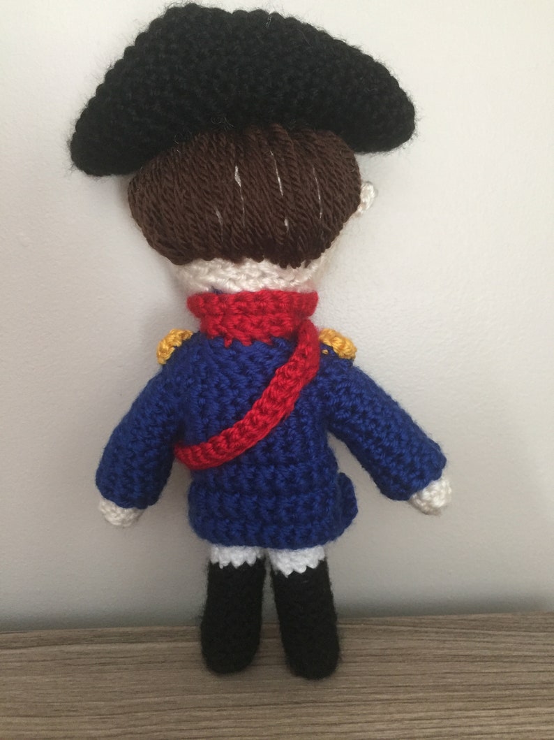May include: A crocheted doll wearing a blue jacket with red and gold accents, a black hat, and brown hair.