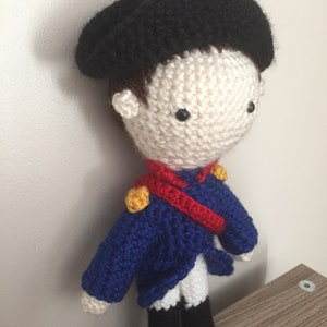 May include: A crocheted doll wearing a black beret, blue jacket, red sash, and white pants. The doll has a white face with black eyes and a brown crocheted hair.