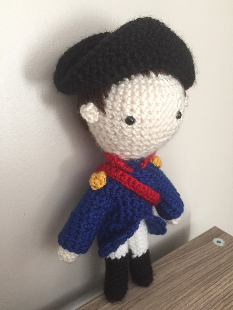 May include: A crocheted doll wearing a black beret, blue jacket, red sash, and white pants. The doll has a white face with black eyes and a brown crocheted hair.