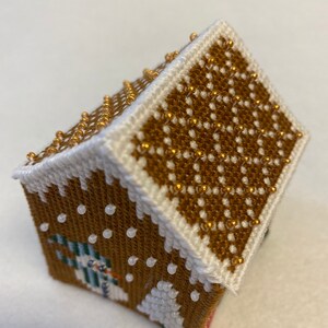 May include: A small gingerbread house cross-stitch with a snowman and a tree on the front. The roof is covered in gold beads and the house is decorated with white beads.