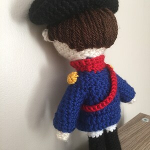 May include: A crocheted doll wearing a blue and red uniform with a black hat. The doll has brown hair and white skin.