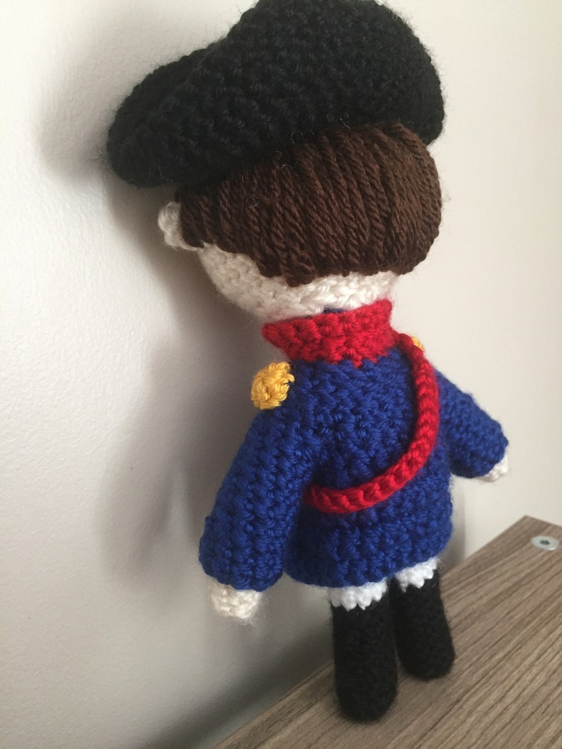 May include: A crocheted doll wearing a blue and red uniform with a black hat. The doll has brown hair and white skin.