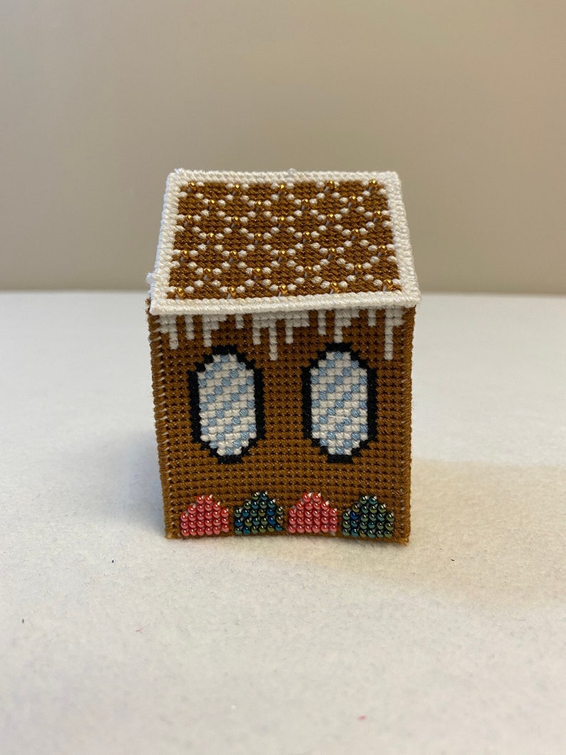 May include: A small gingerbread house made of felt and beads. The house is brown with white icing and blue and white striped windows. The roof is decorated with gold beads. The base of the house is decorated with pink and green beads.