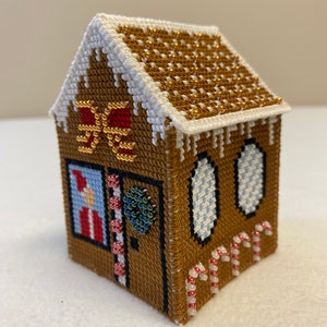 May include: A miniature gingerbread house cross-stitch with a white roof, brown walls, red and white candy canes, and a green beaded wreath on the door.