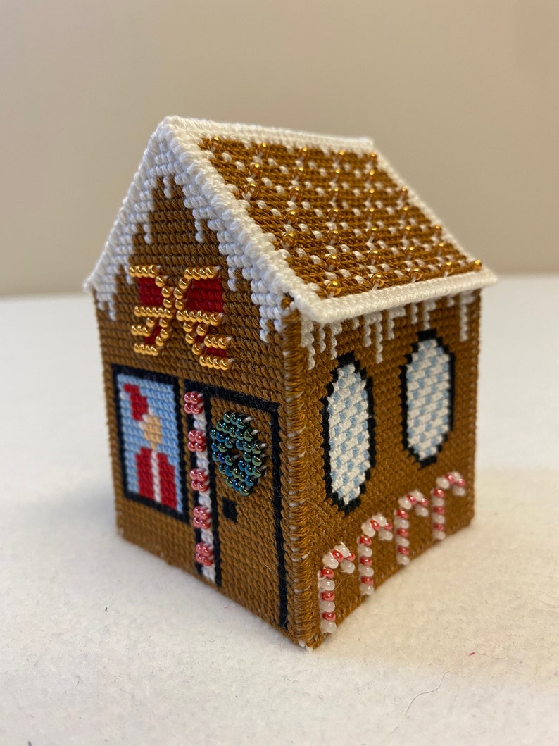 May include: A miniature gingerbread house cross-stitch with a white roof, brown walls, red and white candy canes, and a green beaded wreath on the door.