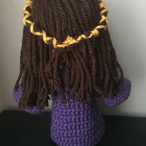 May include: A crocheted doll with long brown hair and a yellow braid. The doll is wearing a purple dress.