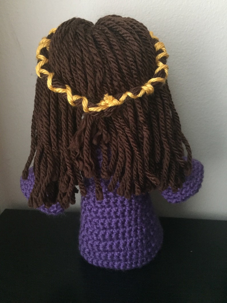 May include: A crocheted doll with long brown hair and a yellow braid. The doll is wearing a purple dress.