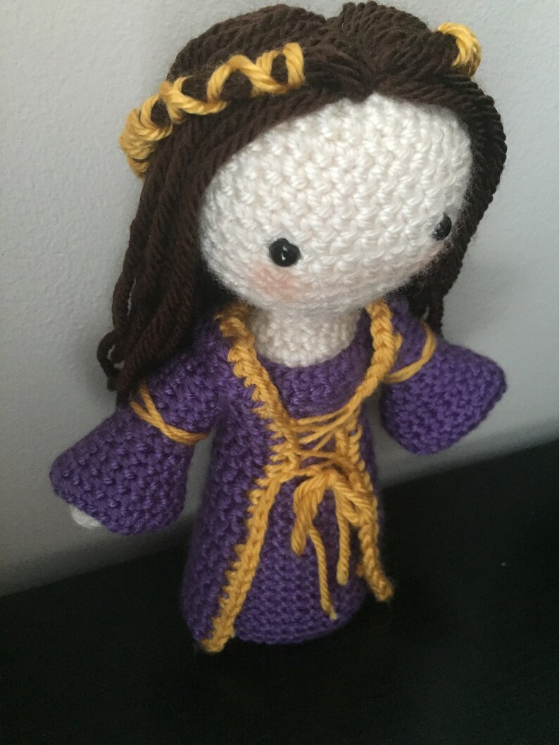 May include: A crocheted doll with brown hair and a purple dress with yellow trim. The doll has a white face with black eyes and a pink blush.