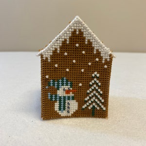 May include: A brown gingerbread house cross-stitch with white snow, a snowman with a blue and green hat and scarf, and a green pine tree.