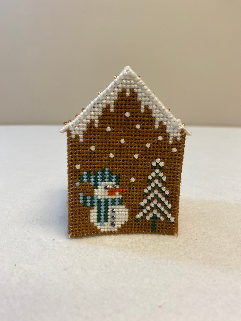 May include: A brown gingerbread house cross-stitch with white snow, a snowman with a blue and green hat and scarf, and a green pine tree.