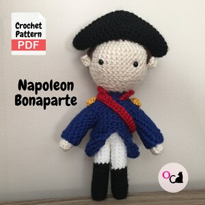 May include: Crochet pattern PDF for a Napoleon Bonaparte amigurumi doll. The doll is wearing a blue jacket with red and yellow accents, a white shirt, and black boots. The doll has a black hat and a white face with black eyes.