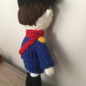 May include: A crocheted doll wearing a blue jacket, red scarf, and black hat. The doll has brown hair and white skin.