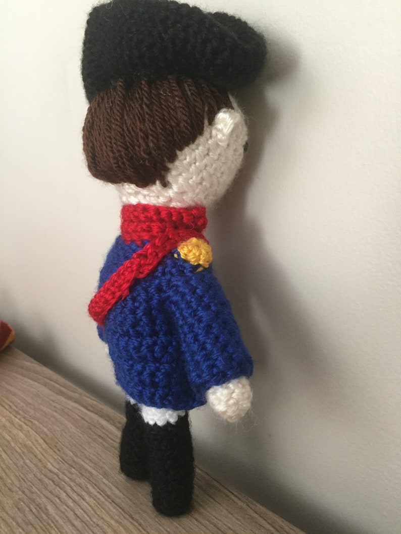 May include: A crocheted doll wearing a blue jacket, red scarf, and black hat. The doll has brown hair and white skin.
