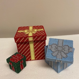 Pode incluir: Três pequenas caixas de presente, uma vermelha com um laço dourado, uma azul com um laço prateado e uma verde com um laço vermelho.