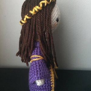 May include: A crocheted doll with long brown hair and a purple dress with yellow trim. The doll is wearing a yellow braid crown.