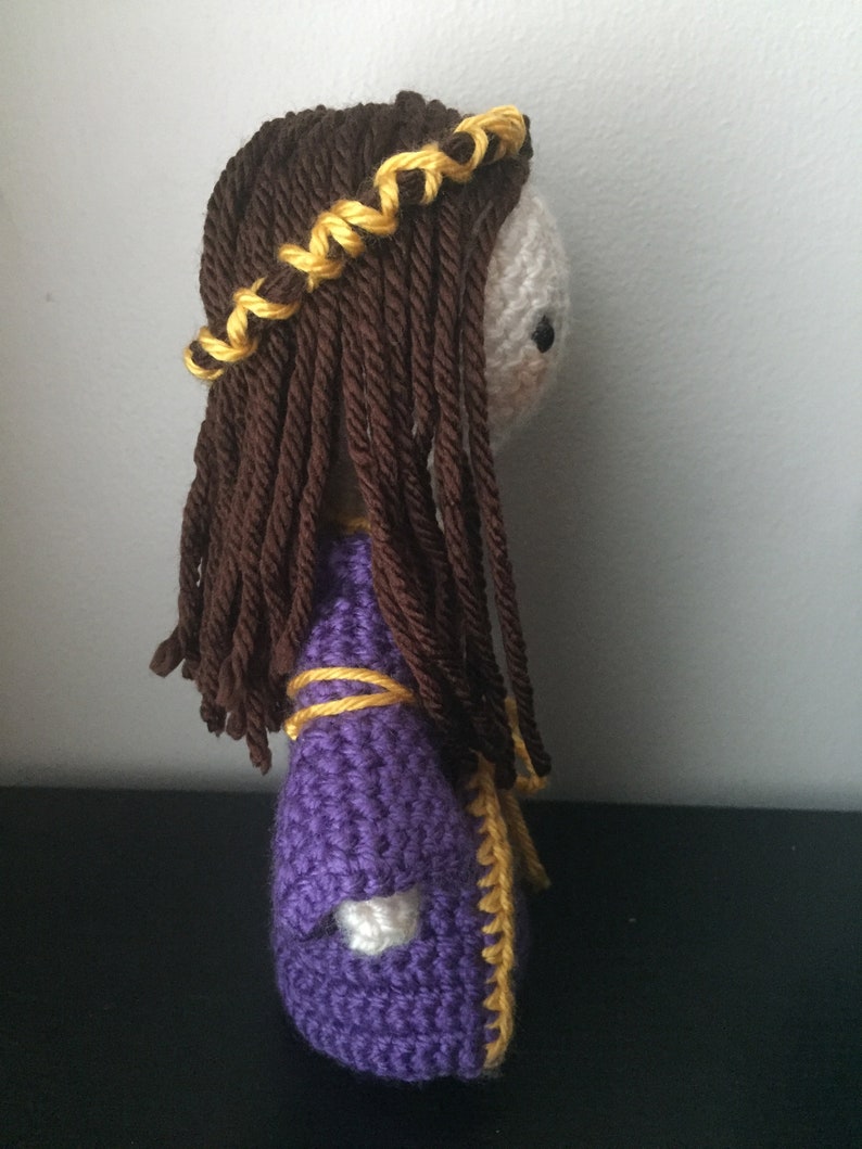 May include: A crocheted doll with long brown hair and a purple dress with yellow trim. The doll is wearing a yellow braid crown.