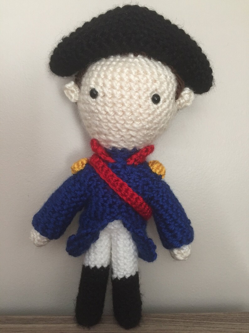 May include: A crocheted doll wearing a blue and red uniform with a black hat. The doll has white skin, black eyes, and a red mouth.