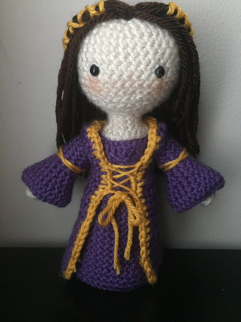 May include: Crocheted doll with brown hair, a purple dress with yellow trim, and a gold crown.