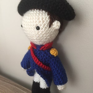 May include: A crocheted doll wearing a blue and white uniform with a red sash and a black hat. The doll has a white face with black eyes and brown hair.