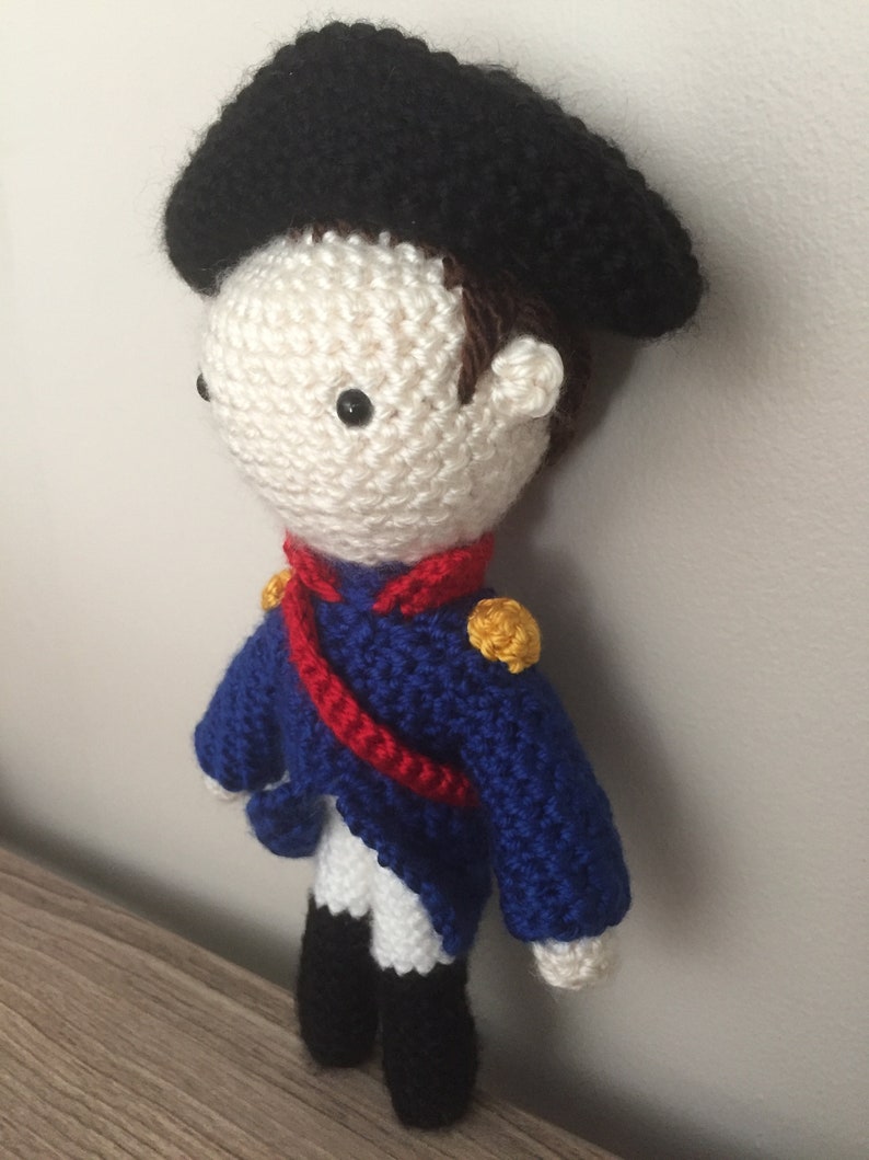 May include: A crocheted doll wearing a blue and white uniform with a red sash and a black hat. The doll has a white face with black eyes and brown hair.