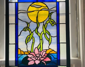 May include: A stained glass window depicting a yellow sun, green bamboo stalks, and a pink water lily in a blue pond. The window is framed in black.
