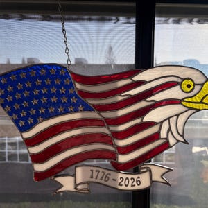 May include: Stained glass American flag and eagle design. The flag features red and white stripes with a blue field of stars. The eagle has a yellow beak and eye. A banner below reads "1776-2026". The piece is suspended by a chain.