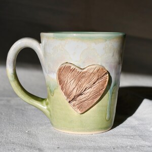 May include: A handmade ceramic mug with a heart-shaped design. The mug features a light green and white glaze with a textured brown heart. The handle and base are green. The mug is a unique piece of drinkware.