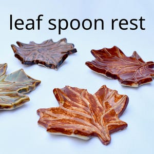 May include: Four ceramic spoon rests shaped like autumn leaves in shades of brown, tan, and green with a glossy finish. The text leaf spoon rest is printed in black above the items. Each piece features detailed embossed veins and irregular edges.