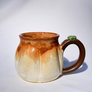 May include: A ceramic mug with a rounded, fluted body featuring a gradient glaze that transitions from a deep brown rim to a creamy beige base. The mug has a dark brown handle topped with a small, glossy green accent piece at the attachment point.