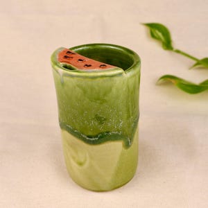 Pode incluir: Um copo cilíndrico de cerâmica com um esmalte verde brilhante, apresentando um motivo decorativo de fatia de melancia na borda. O recipiente tem um corpo liso e arredondado com uma sutil variação de cor do verde claro ao verde escuro.