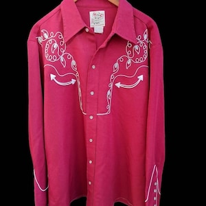 Op de afbeelding: Een lang mouwig, roze westernhemd met witte borduursels. Het shirt heeft een puntige kraag, een knoopsluiting en een enkele borstzak. De borduursels tonen een bloemmotief en een juk.