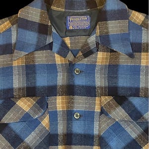 Può includere: Una camicia a quadri blu, grigia e marrone con due tasche sul petto. La camicia ha un colletto e un'etichetta che recita "Pendleton Woolen Mills". La camicia è realizzata in misto lana.